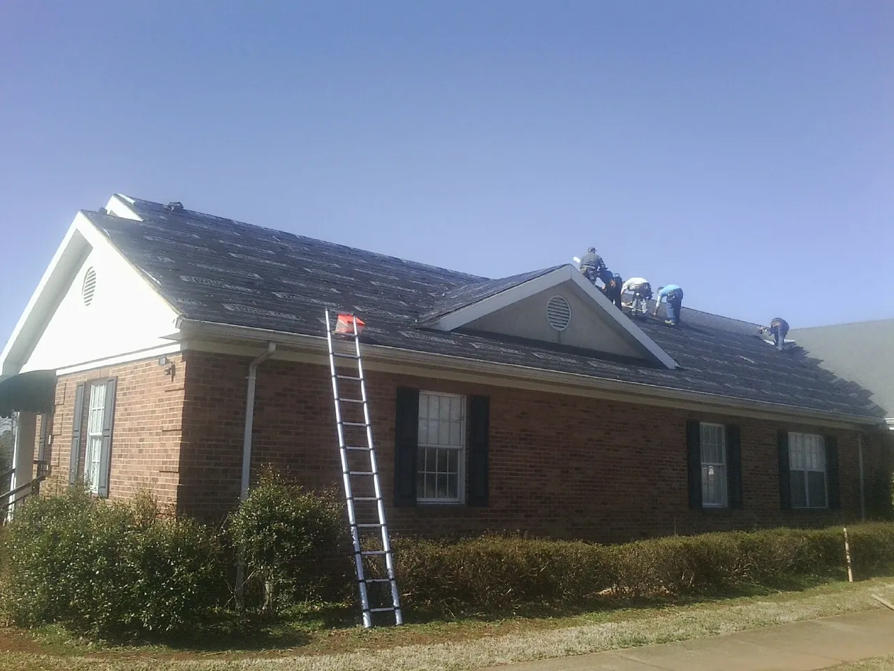 Crew tearing shingles off a brick ranch roof with a ladder set — Emergency Roof Repair work by our crew in Pine Valley